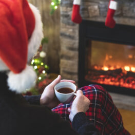 A person wearing a Santa hat sits alone by a glowing fireplace, holding a warm mug, capturing the quiet emotions of spending the holidays in a separated household.