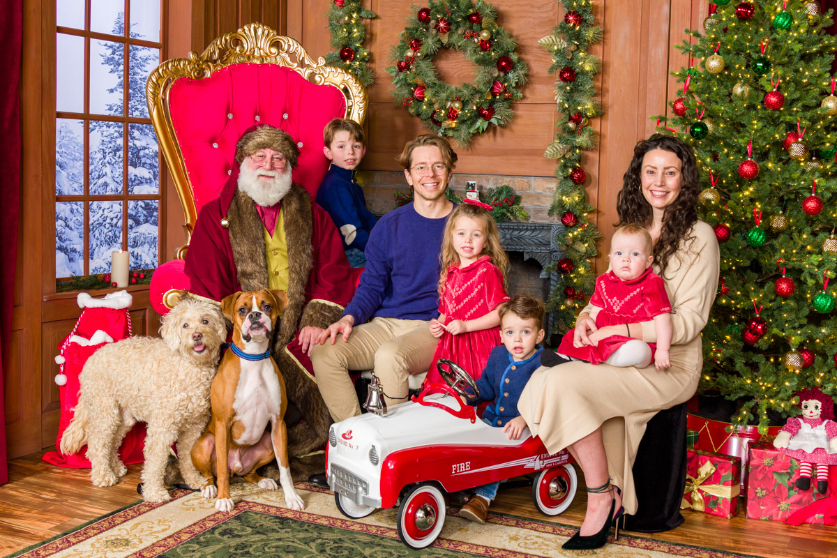 A family with four children and two dogs pose for a holiday photo with Santa, all smiling together in front of festive decorations.