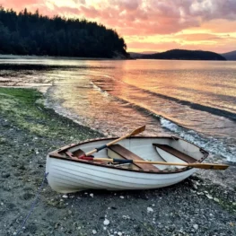 Row boat is moored on the beach, with a gorgeous orange and yellow sunset behind it against the water.