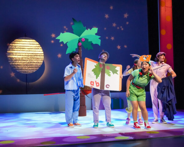Actors gather around a giant storybook prop and coconut tree on stage in Chicka Chicka Boom Boom: The Musical at Seattle Children’s Theatre.