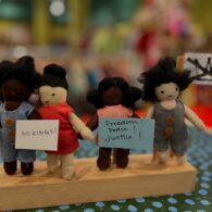 Four handmade cloth dolls stand side by side holding miniature protest signs reading “NO KINGS!” and “Freedom! Peace! Justice!” in front of a colorful background at Chrysanthemum, a children’s store in Seattle.
